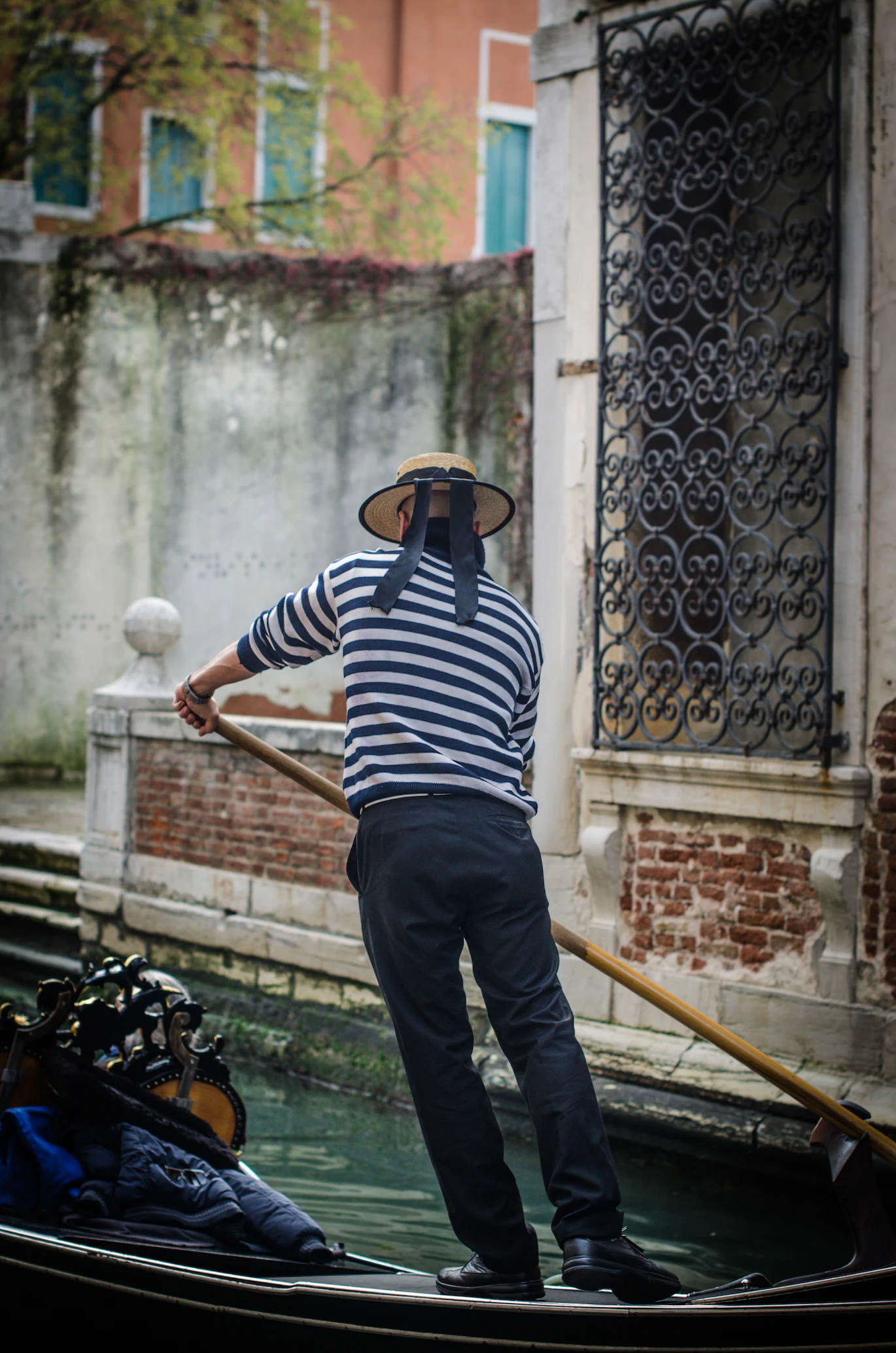 Gondolier in a straw hat and striped shirt seen from behind, poling beside an ornate iron-grilled window