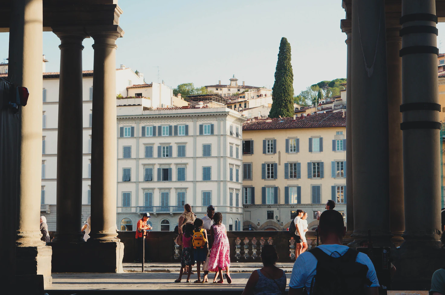 Children walking between stone columns of a loggia, with Florentine palazzi and a cypress behind