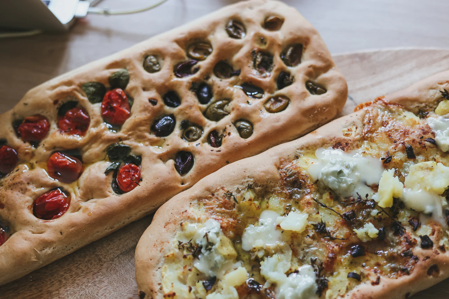 Two rustic focaccias, one topped with cherry tomatoes and olives, the other with cheese and herbs