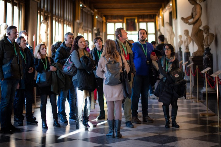 Tour group with audio guides listening to their guide in a sunlit Uffizi gallery with marble statues