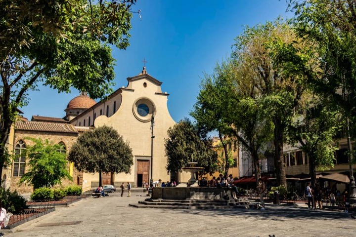 Sunny square in Florence with a plain-faced church, a fountain and people sitting in the shade of trees