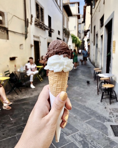 Hand holding a two-scoop gelato cone in a narrow Florentine street with cafe tables