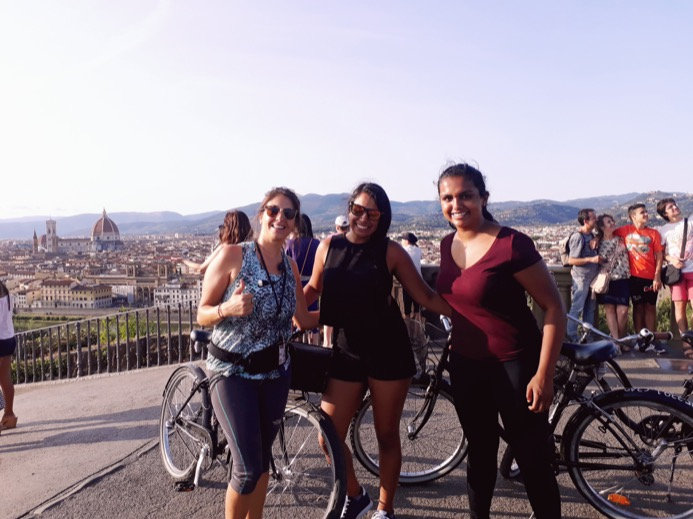 Three smiling travellers with their bikes at a hilltop viewpoint over Florence