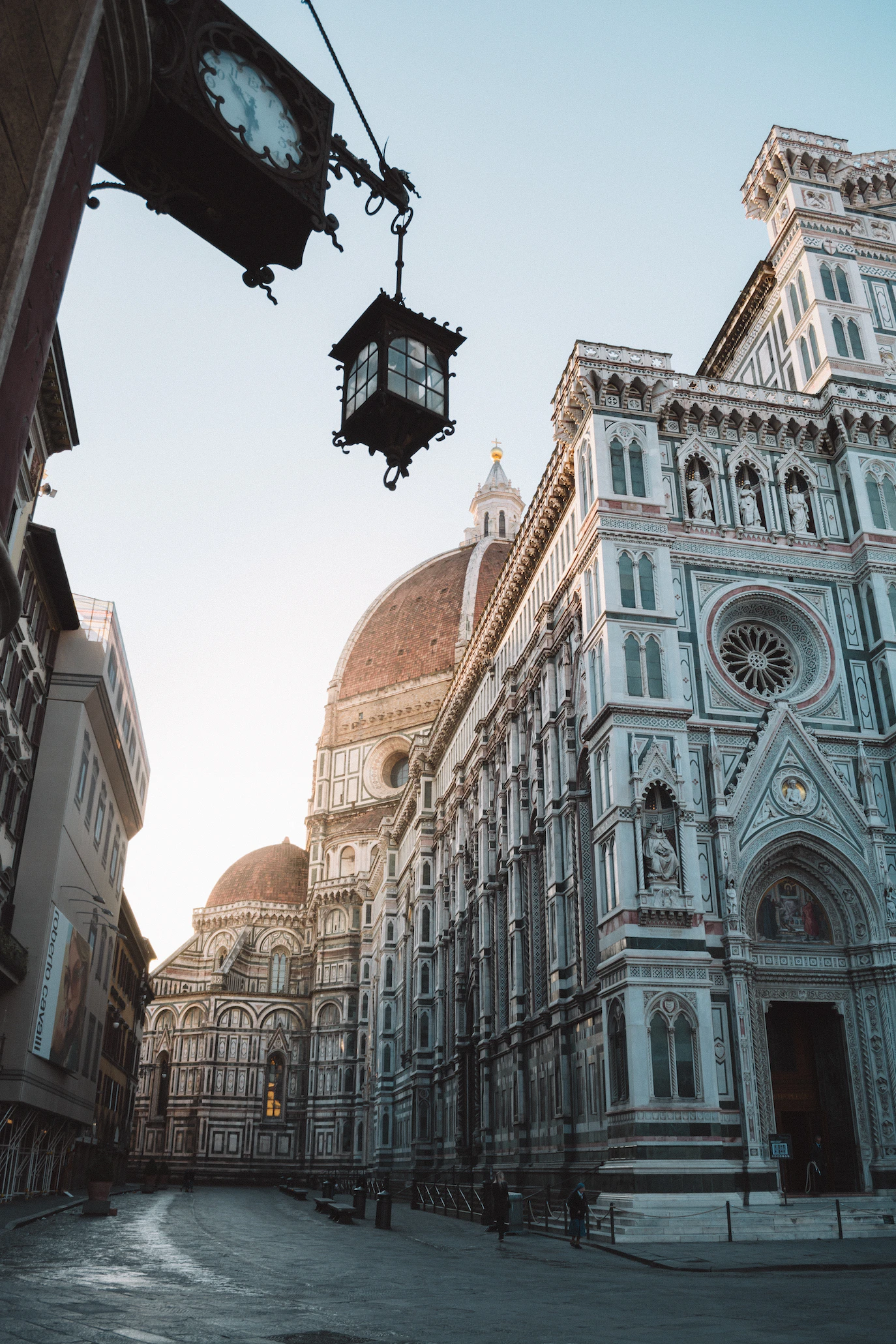Quiet Florence street at dawn with the cathedral's marble facade and dome beyond a hanging street lamp