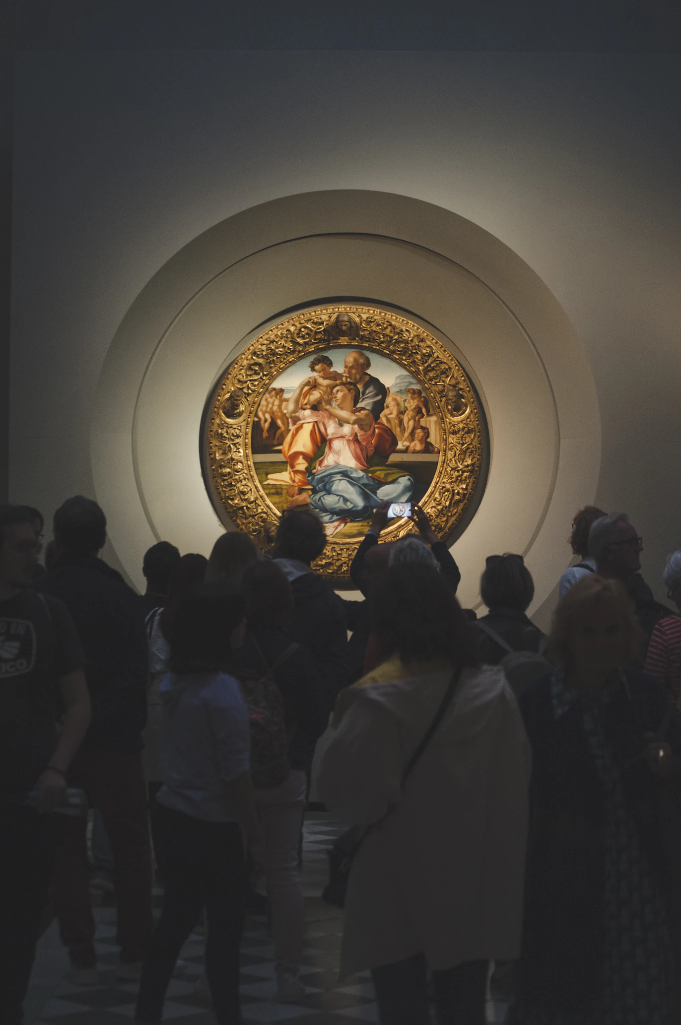 Visitors crowding in a darkened gallery around a circular Renaissance painting in a golden frame