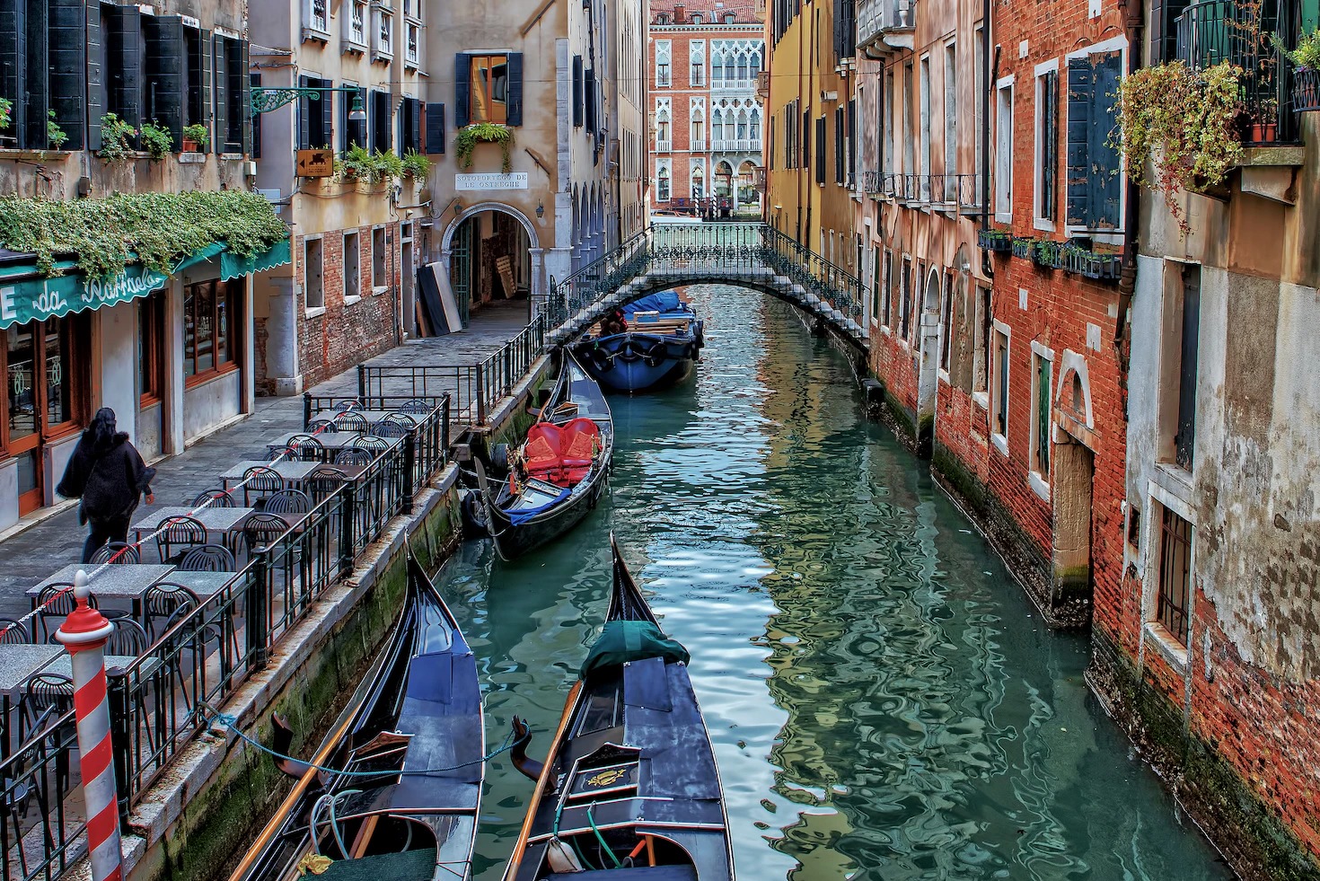 Gondolas moored on a green Venetian canal beside a café terrace and a small bridge