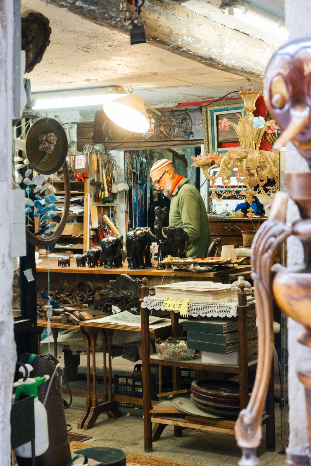 Craftsman at work in a cluttered Venetian workshop filled with carved wood and chandeliers