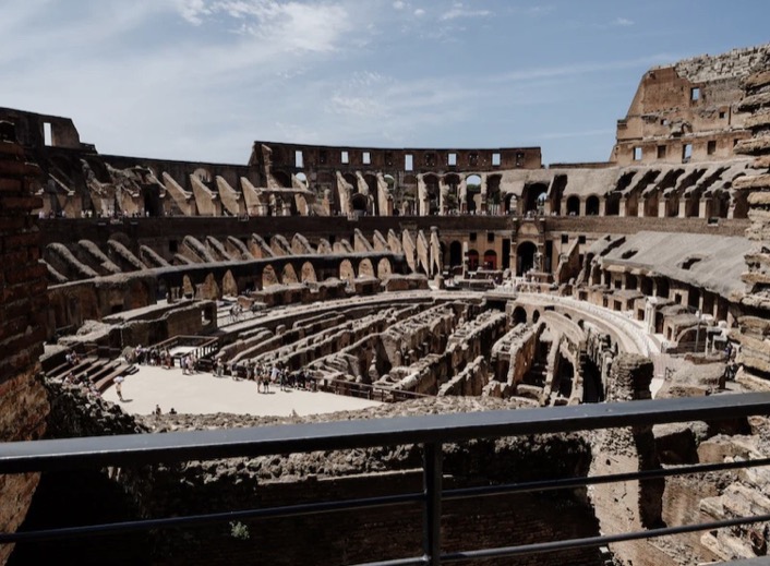 Interior of the Colosseum with the arena floor and tiers of seating seen from a railed balcony