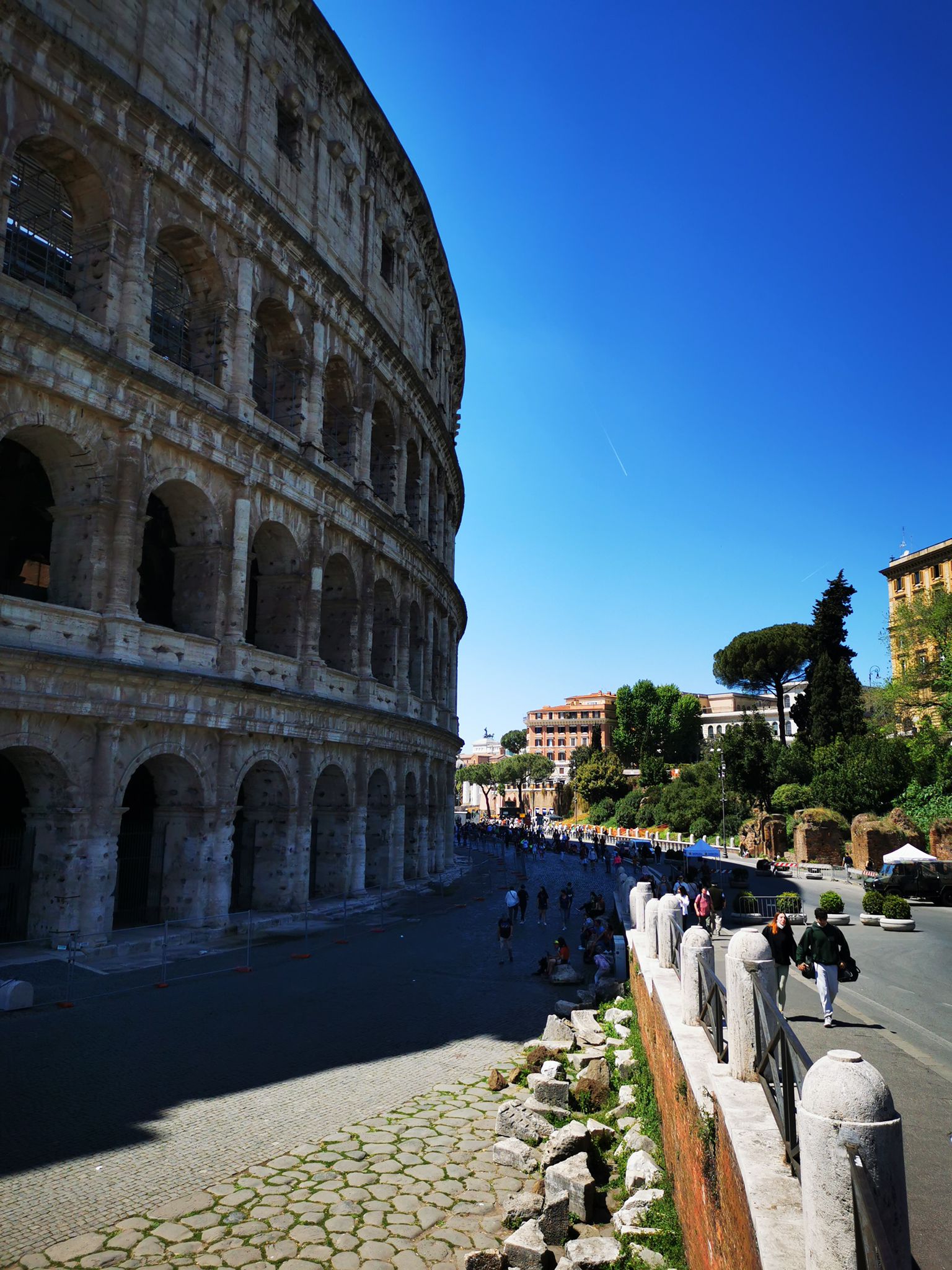 Curving arcaded wall of the Colosseum along a cobbled street under a clear blue sky