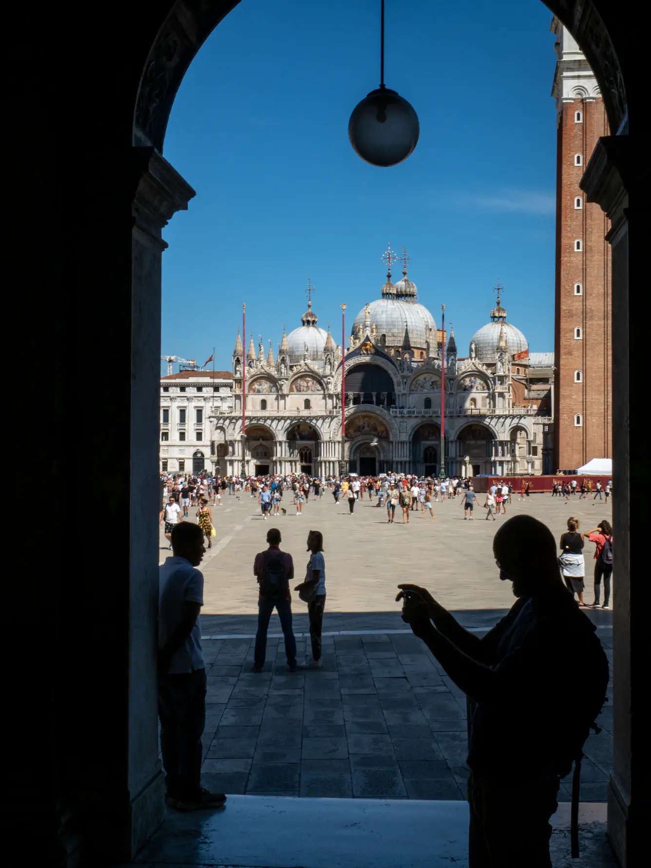 Silhouetted visitors under an arcade looking out onto St Mark's Square and the domes of the Basilica