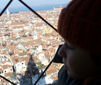 Child in a woollen hat gazing through the bars of a bell tower over the red rooftops of Venice