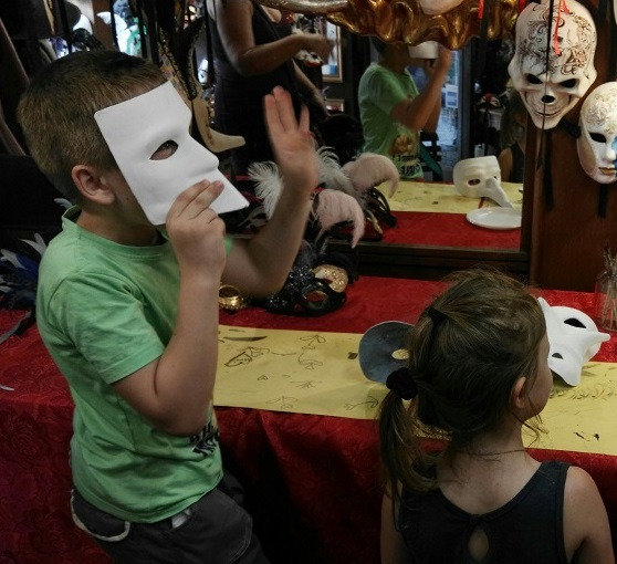 Children trying on white Venetian masks in a mask workshop, with decorated masks hanging on the wall