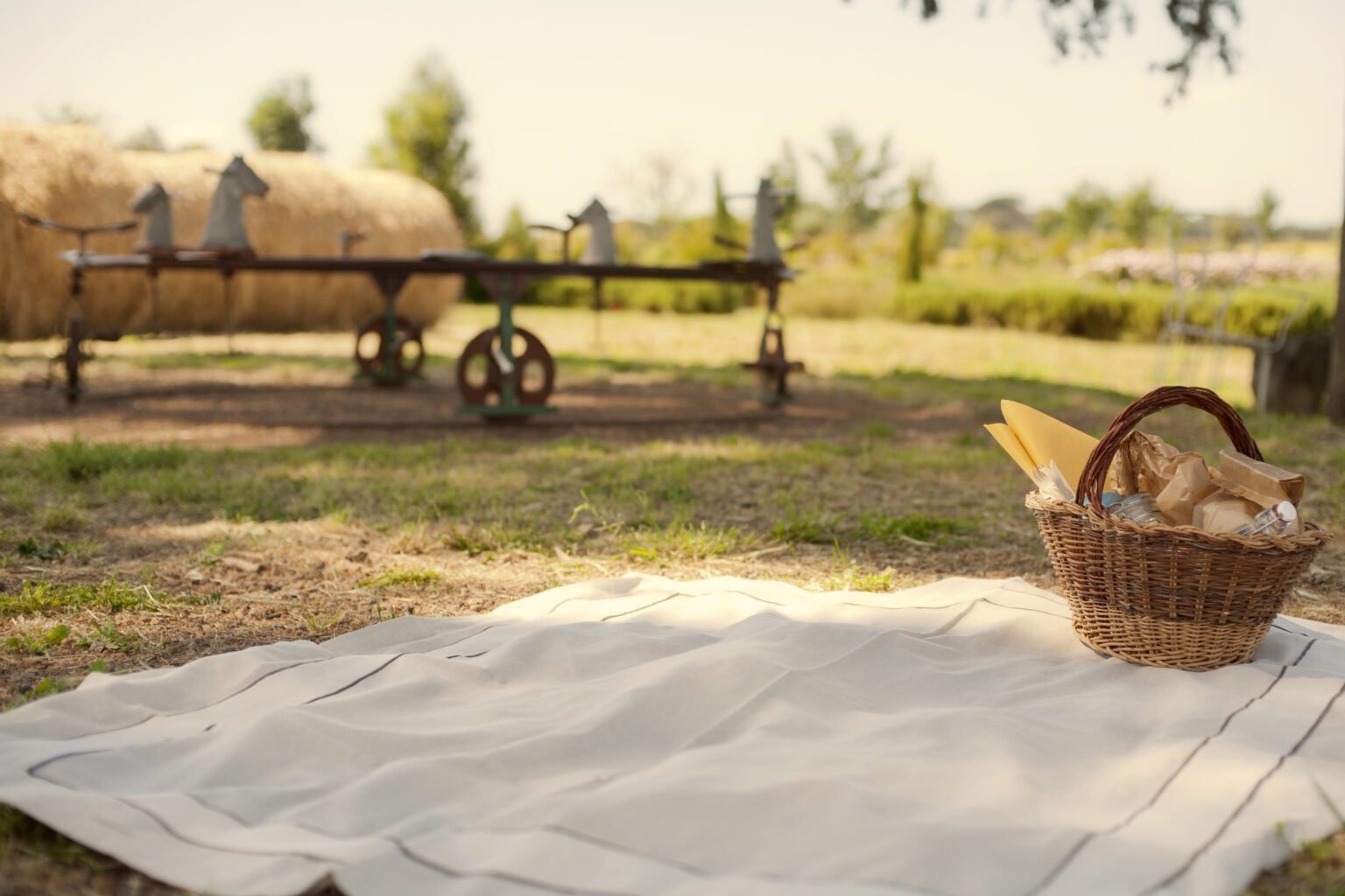 Picnic blanket and a wicker basket of bread on the grass, with hay bales and old toy horses behind