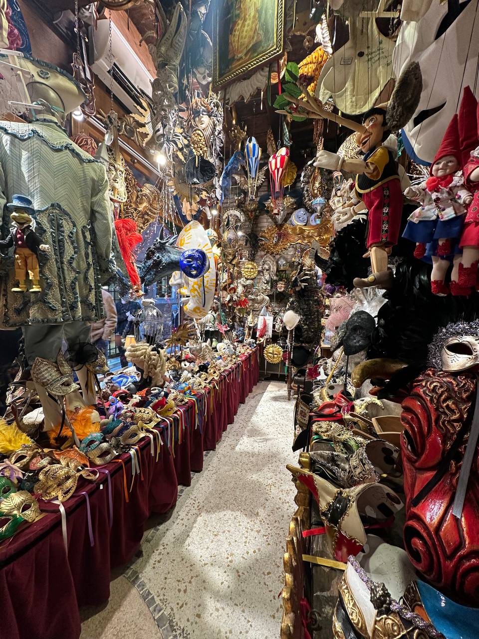 Narrow aisle of a mask shop with velvet-draped tables, puppets and masks on every wall