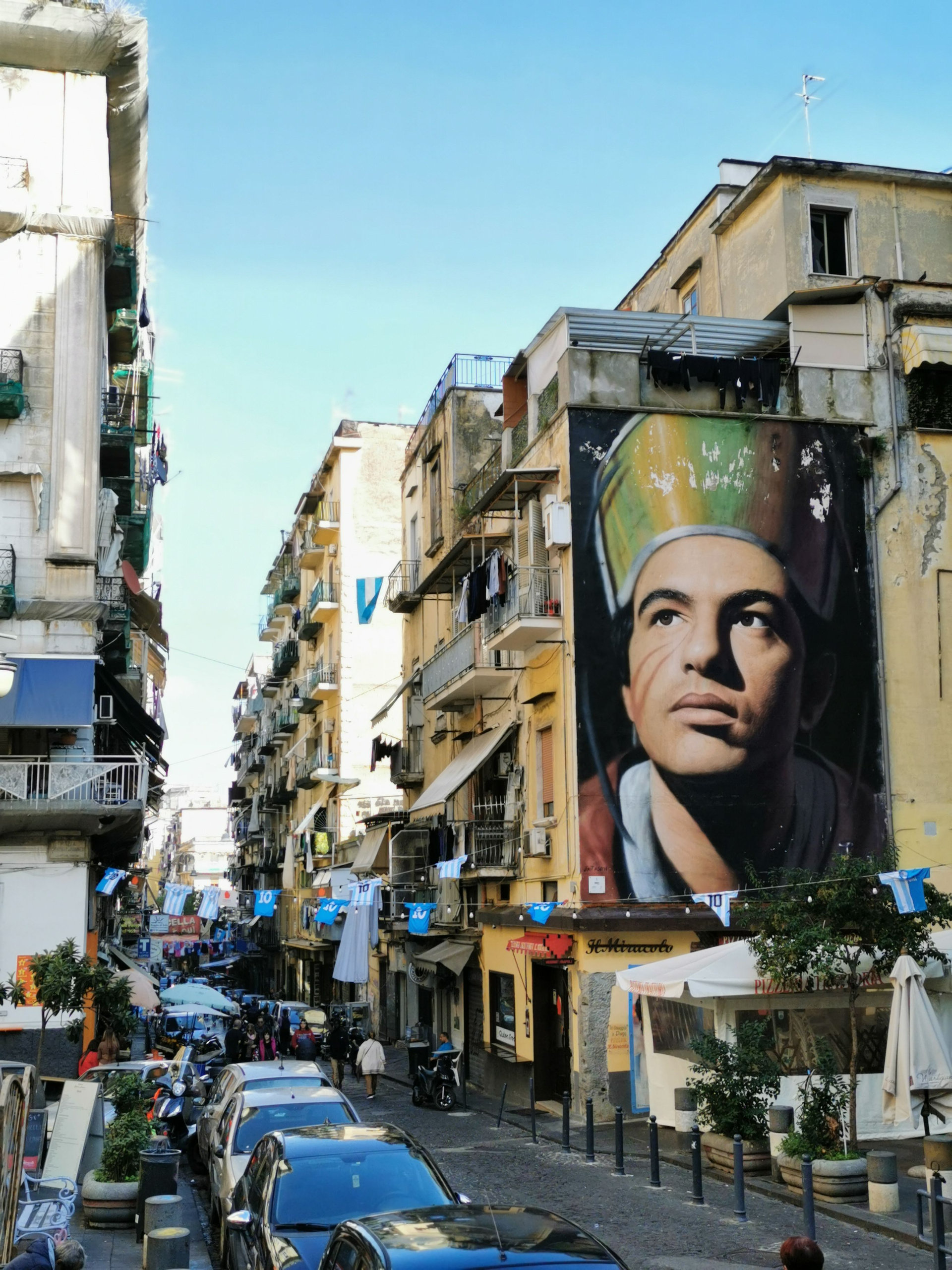 Narrow Naples street lined with tall buildings and a large mural of a bishop's face on a gable wall