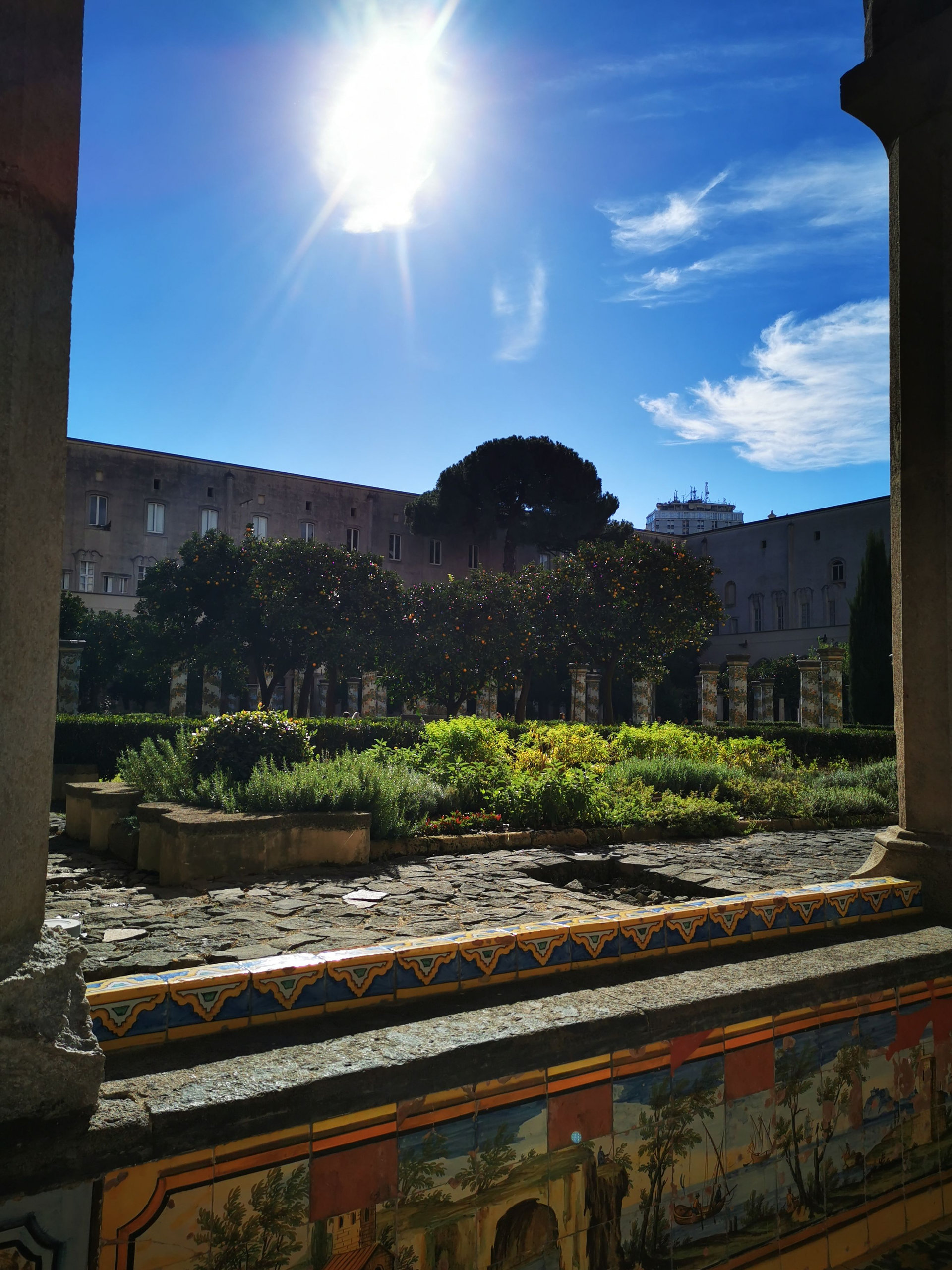 Sunlit cloister garden seen between columns, with a tiled majolica bench in the foreground