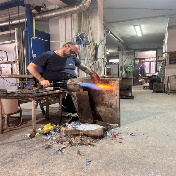 Glassmaker shaping molten glass with a flame in a workshop strewn with coloured glass fragments