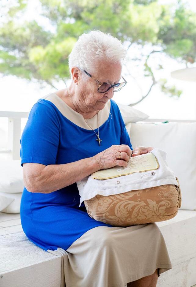 Elderly lacemaker working needle lace on a cushion while seated on a sunny terrace