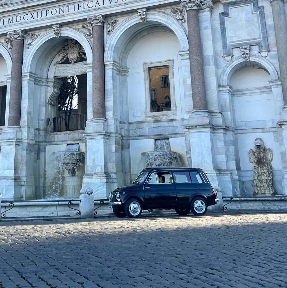 Vintage black car parked before a grand marble fountain with columns and an inscription