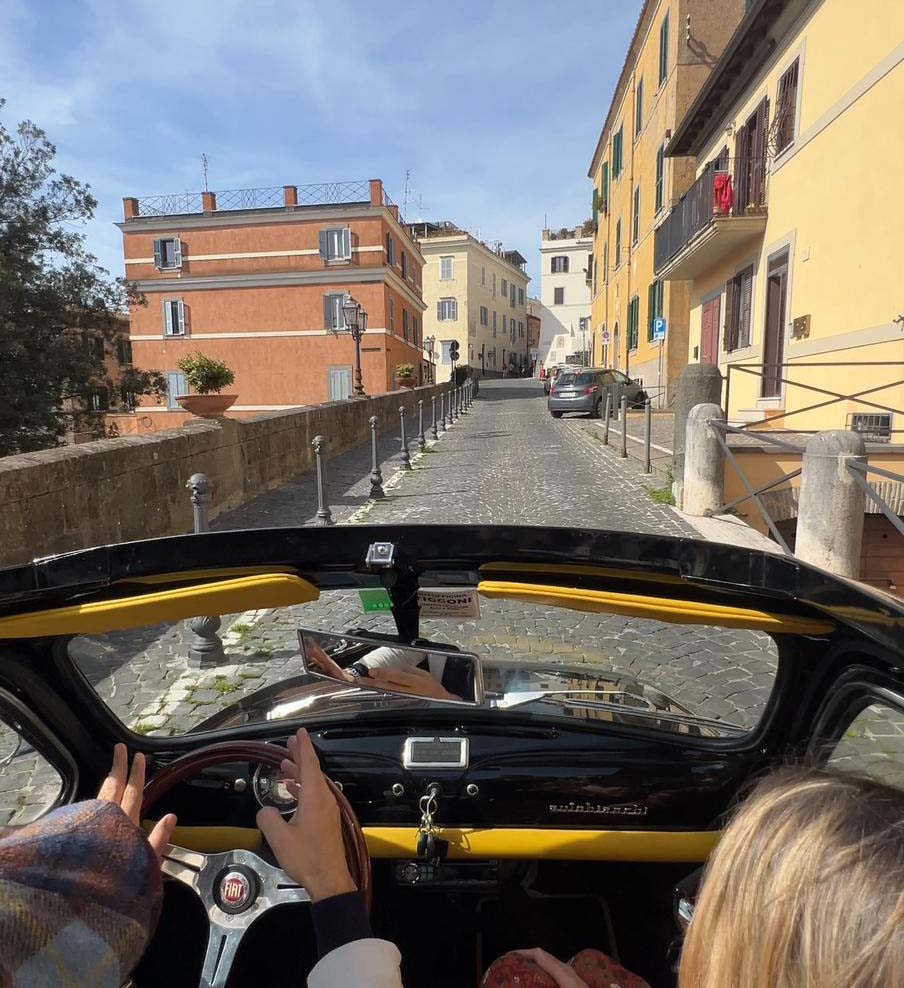 View over the dashboard of a vintage convertible along a cobbled street between ochre houses