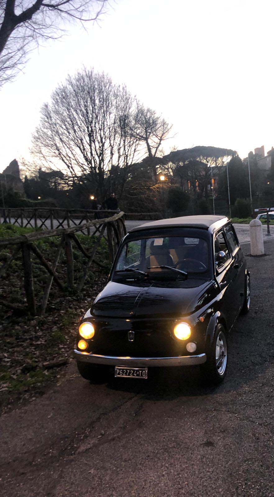 Vintage black Fiat 500 with headlights on at dusk beside a wooden fence and bare trees