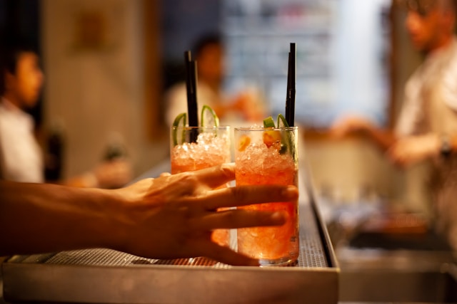 Two icy red spritz cocktails with straws being handed over a bar counter