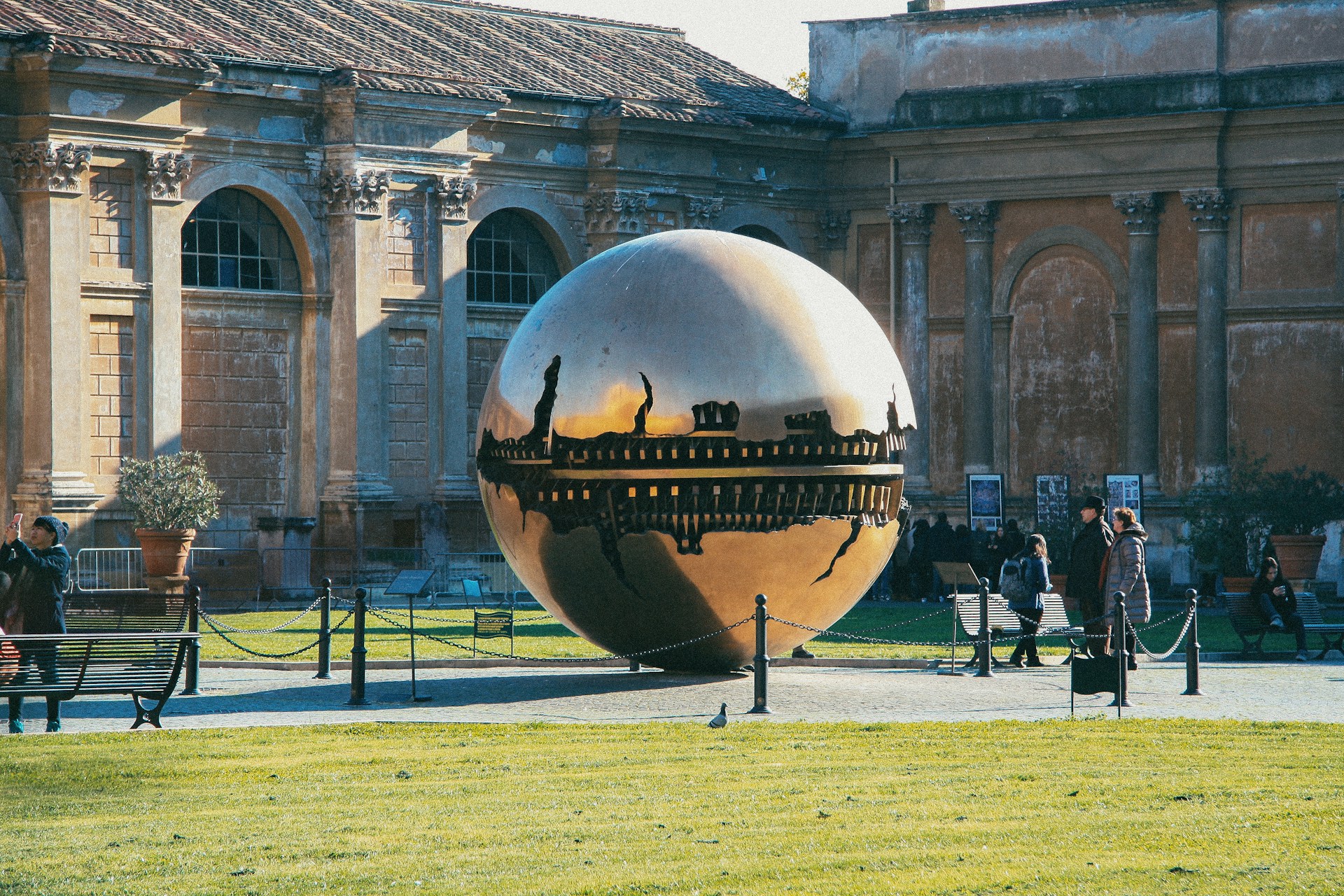Bronze sphere sculpture in a sunlit courtyard of the Vatican Museums with visitors beside it