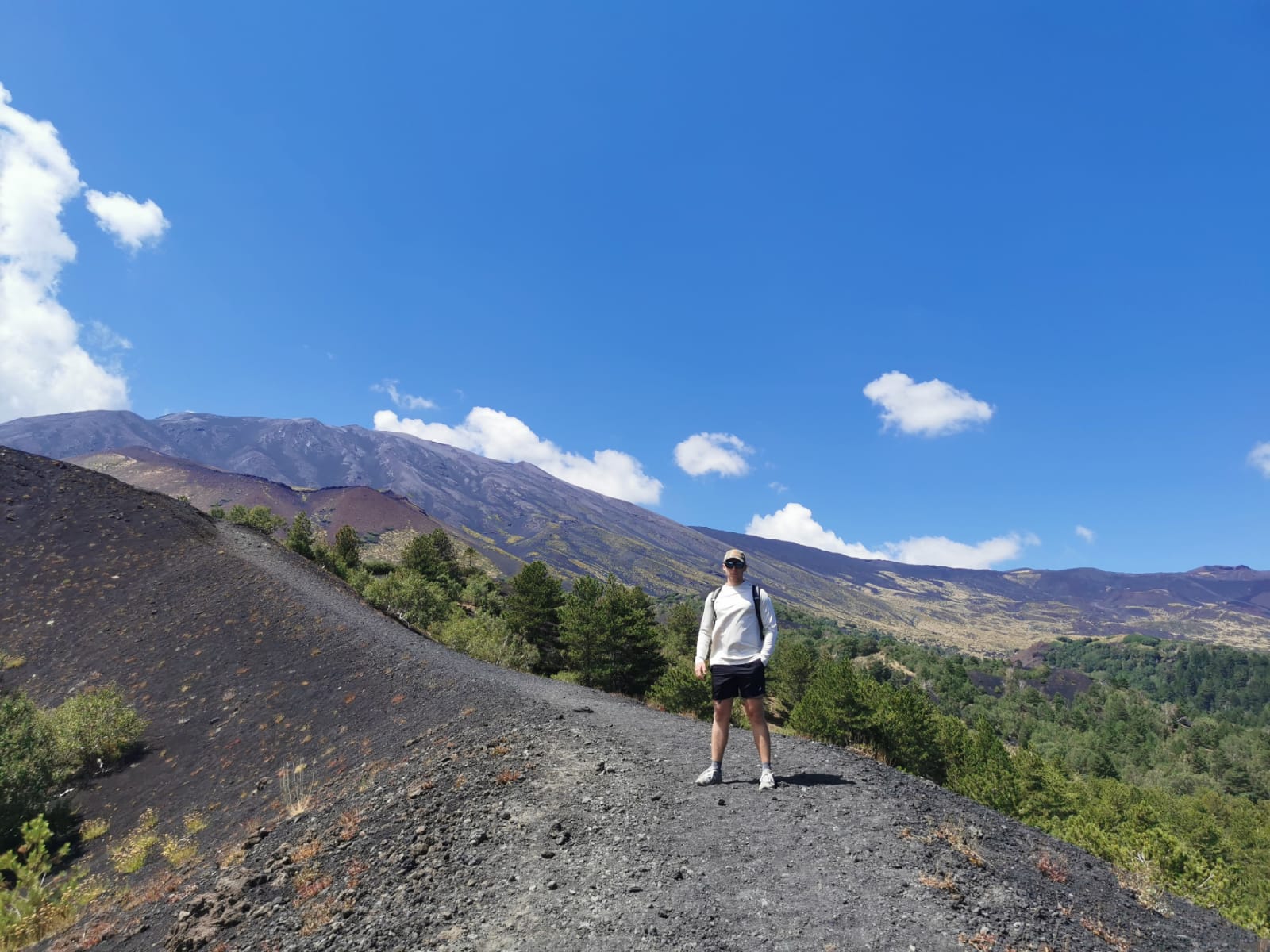 Hiker standing on a dark volcanic ridge of Mount Etna with pine forest and the summit behind