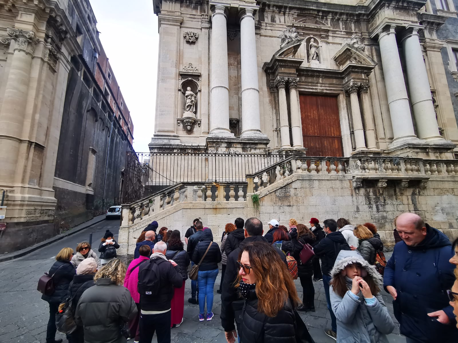 A group of travellers gathered on a street before the Baroque church façade of a Catania basilica
