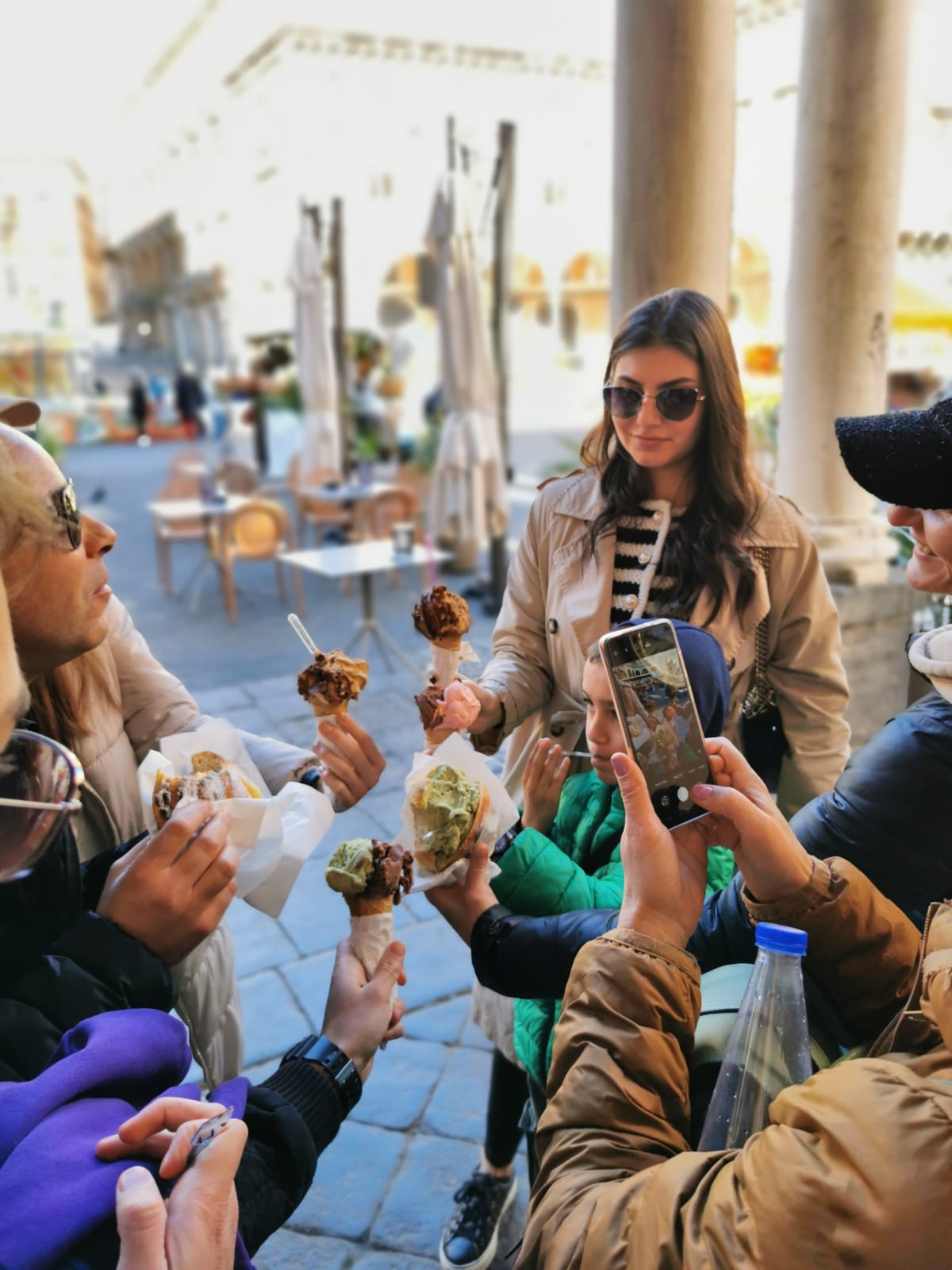 Group of travellers enjoying gelato cones in a sunlit arcade, one taking a photo