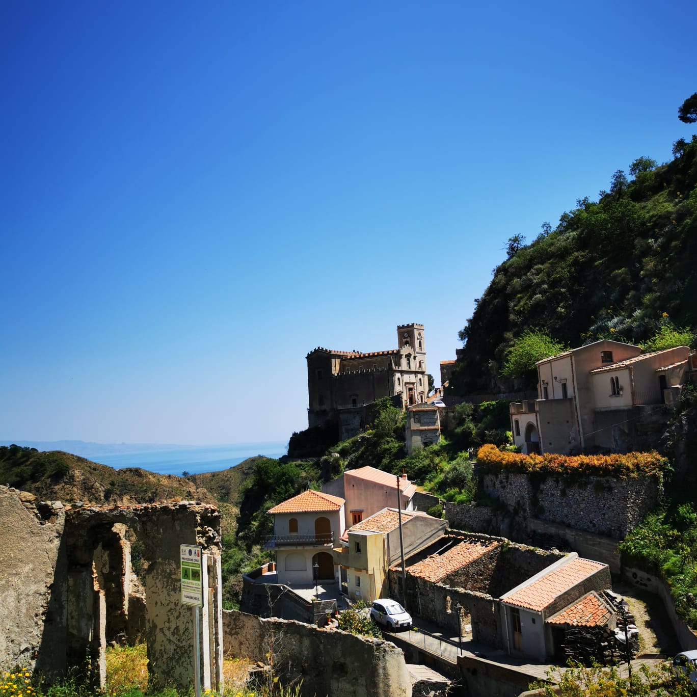 Hillside Sicilian village with a castle-like church tower above ruined walls and a distant sea