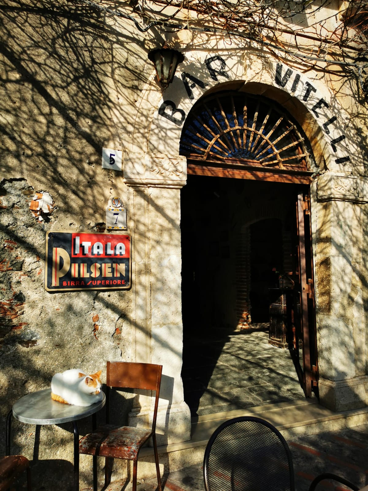 Weathered stone doorway of Bar Vitelli with a vintage beer sign and a cat resting on a cafe table