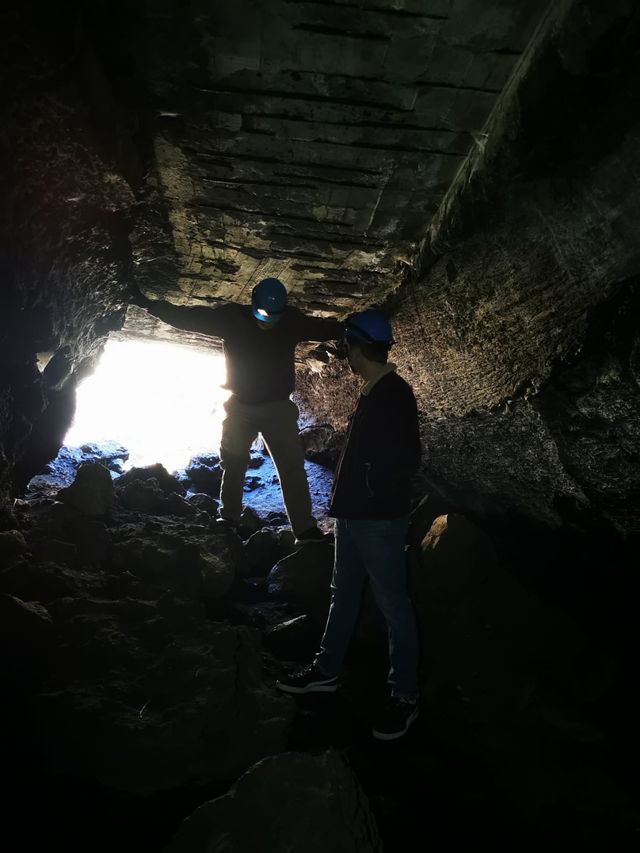 Two people in helmets inside a lava cave, lit by daylight from the entrance