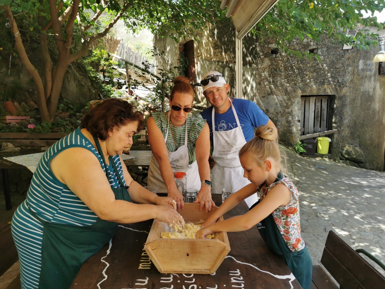 Family and a local cook shaping dough together at an outdoor table in the sun