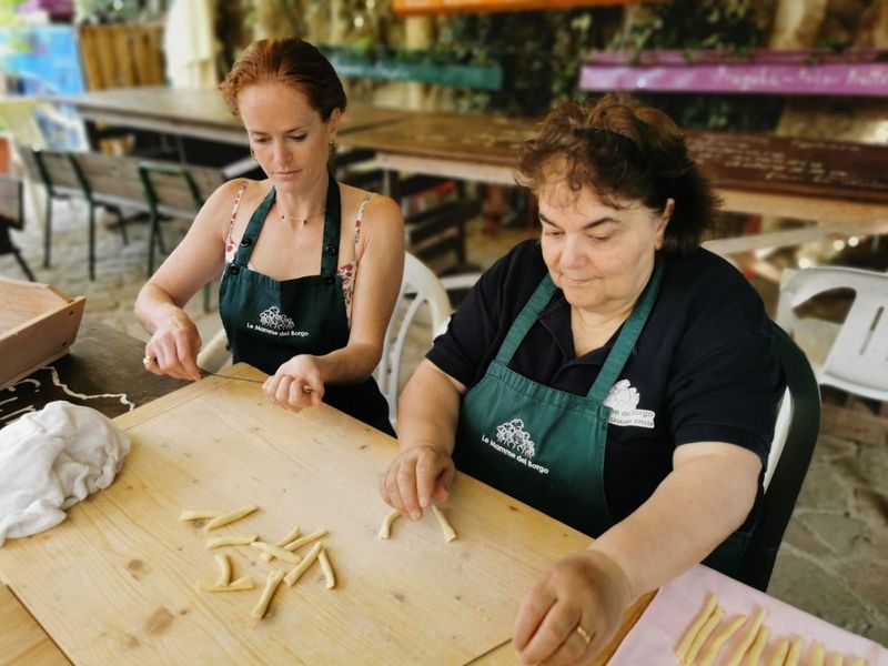 Two women in green aprons rolling small pasta shapes on a wooden board