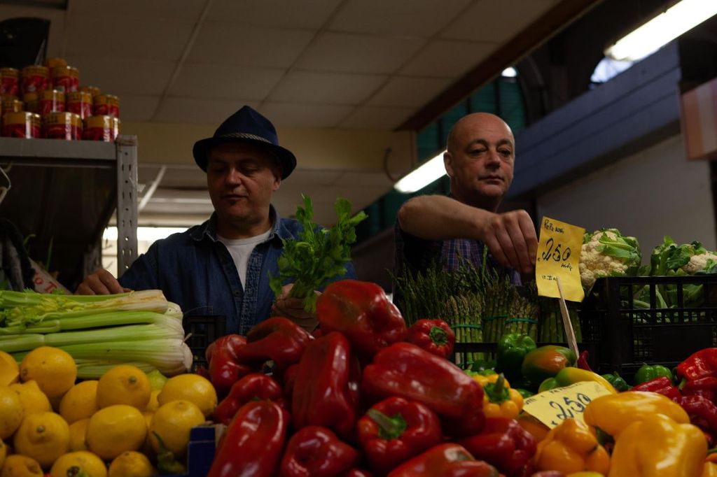Two men browsing a market stall of peppers, lemons and celery
