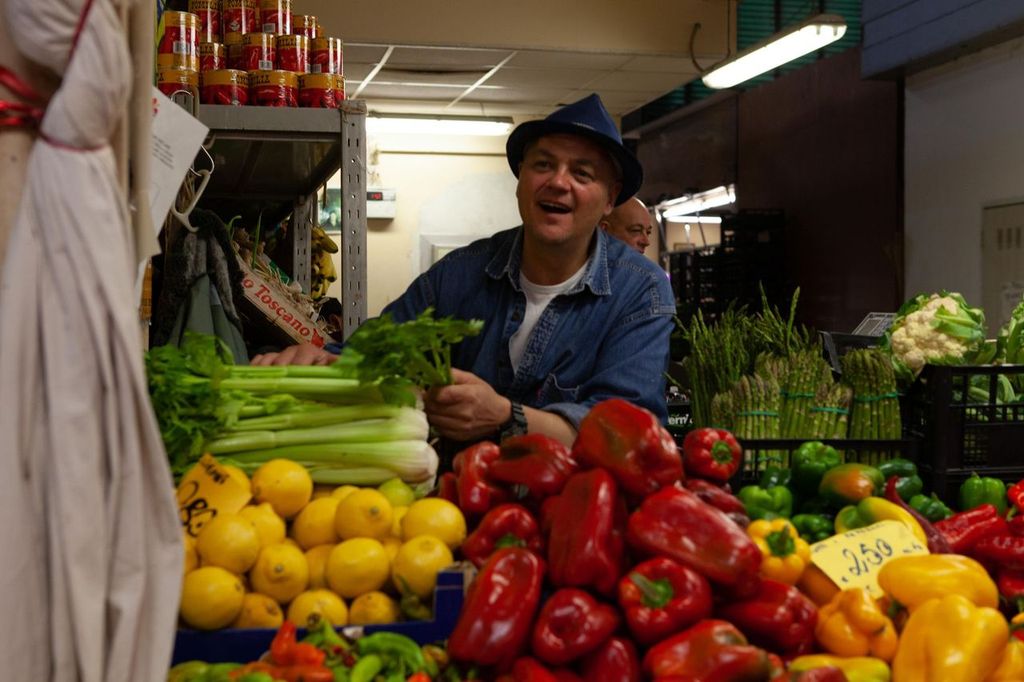 Smiling stallholder in a hat holding celery above crates of peppers and lemons at a market
