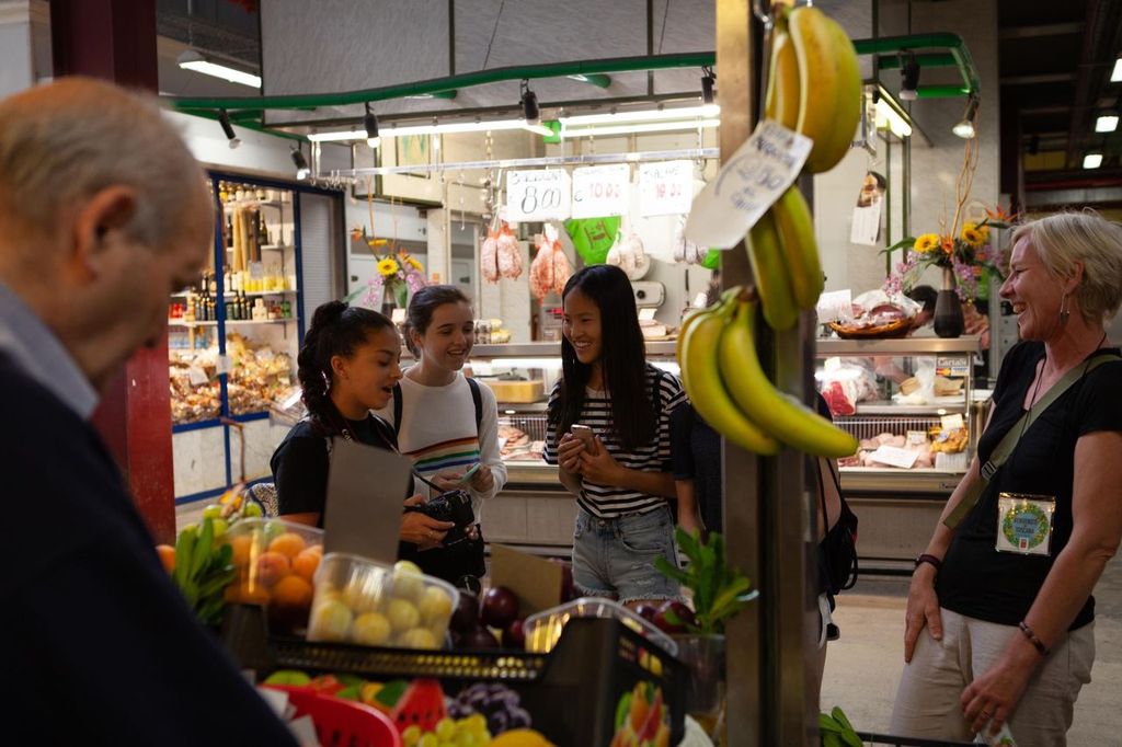 Visitors chatting at a covered market stall hung with bananas and stocked with fruit