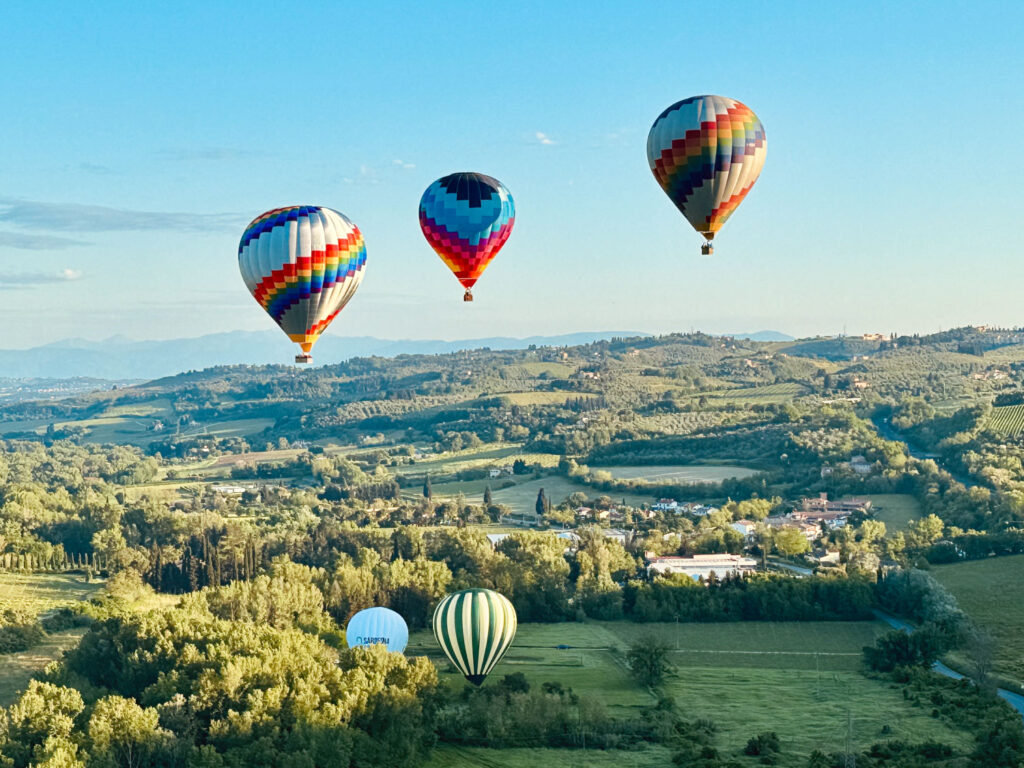 Four colourful hot air balloons floating above rolling Tuscan hills and woodland