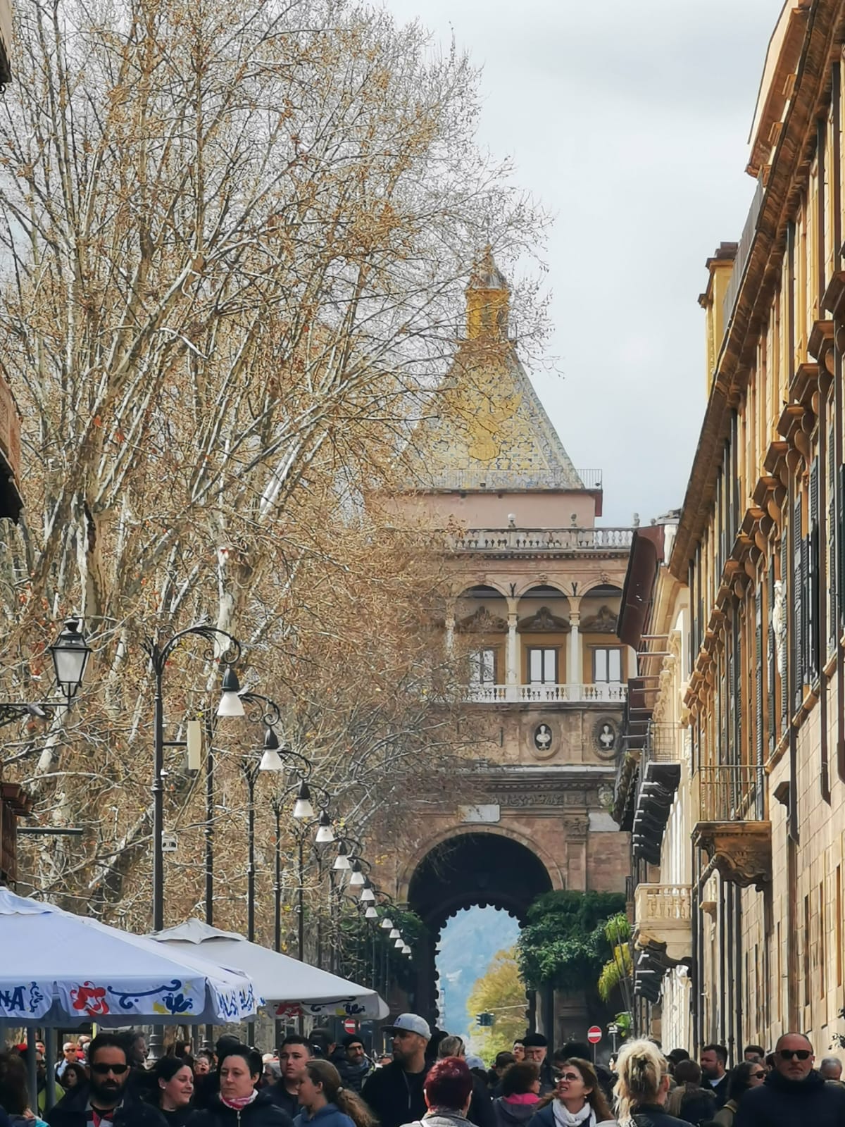 Crowded street lined with plane trees leading toward a monumental gateway with a pyramid-shaped roof