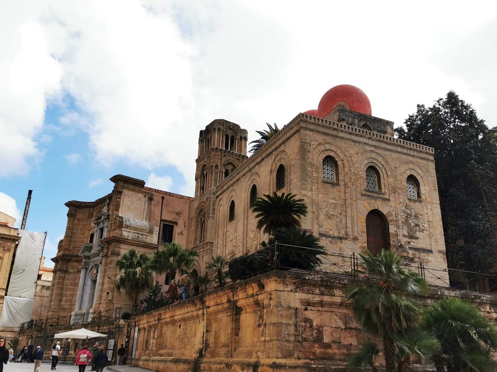 Sandstone church with red domes and a bell tower surrounded by palm trees