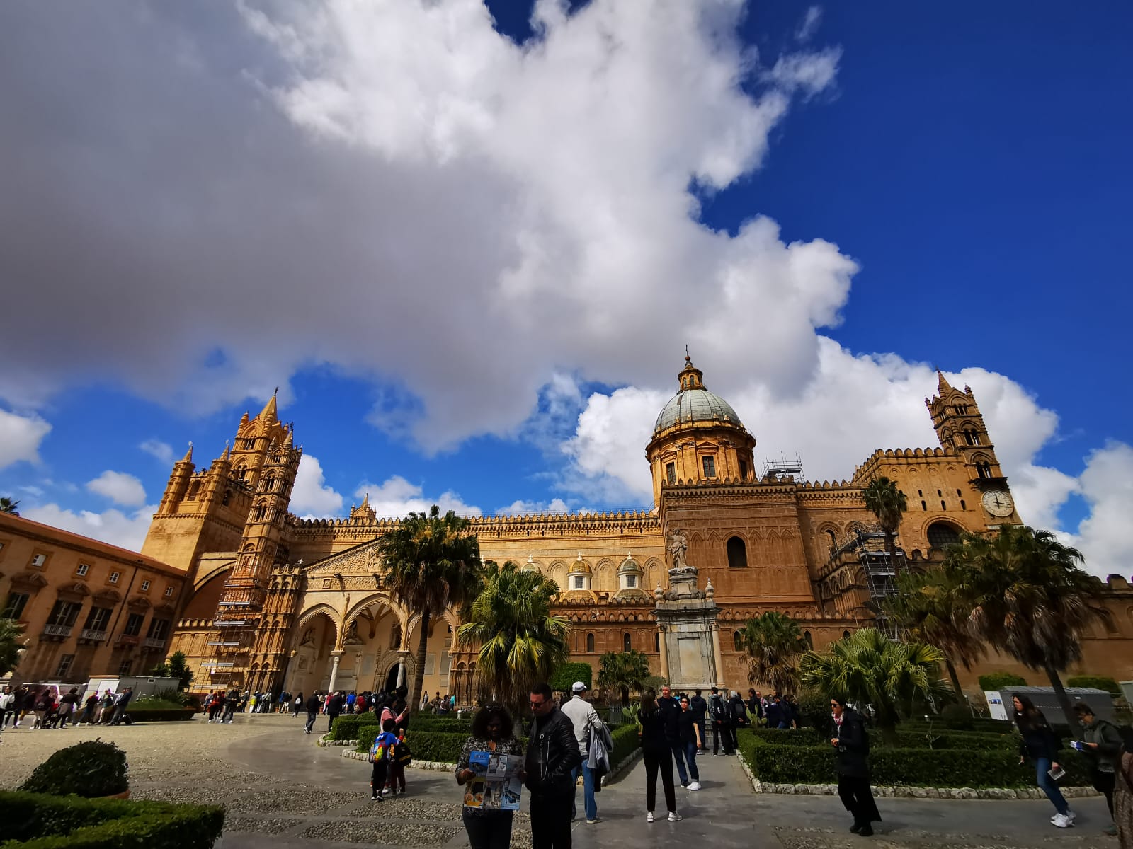 Ornate golden-stone cathedral with towers and a dome rising above a busy piazza and palm trees