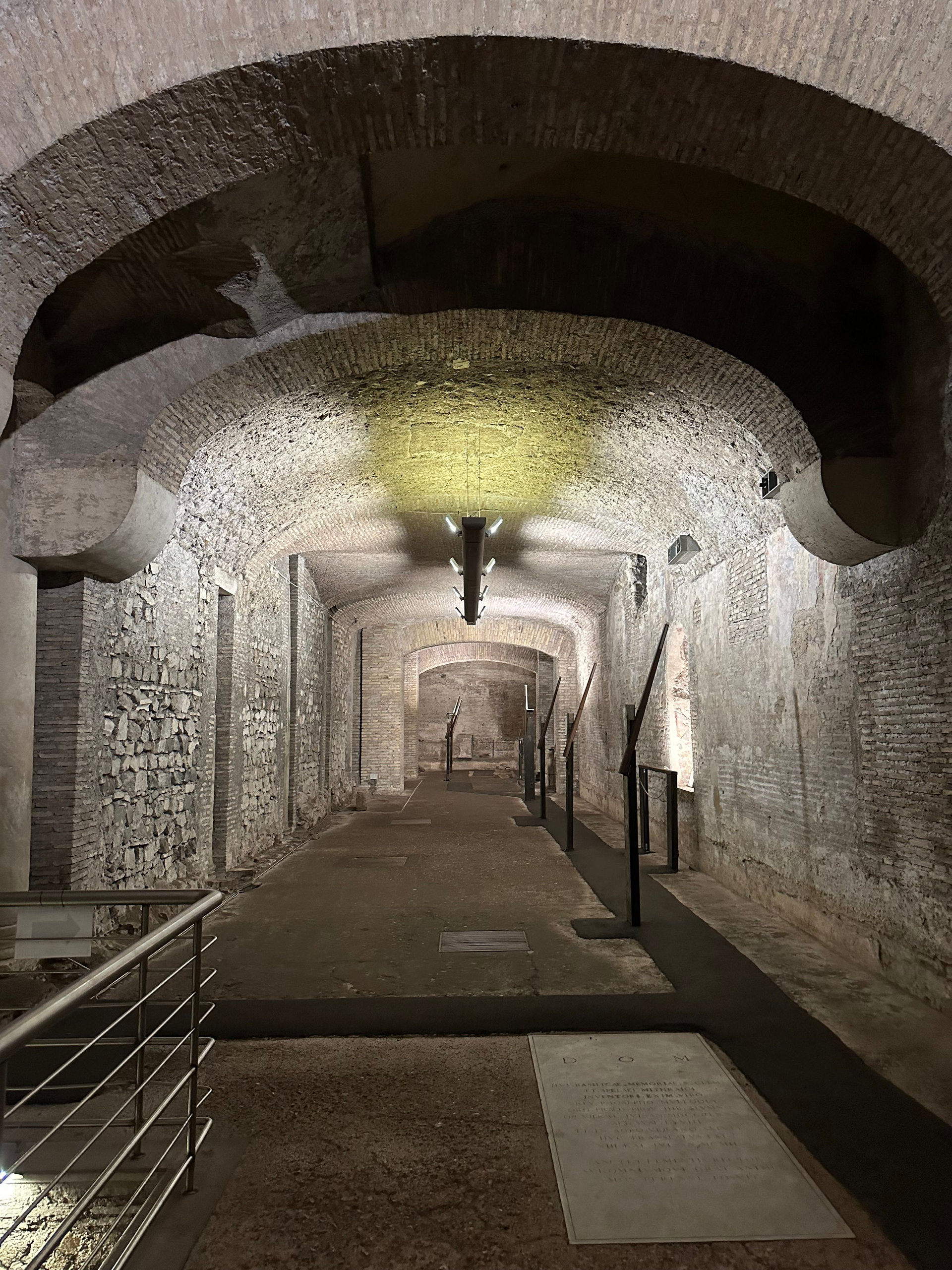 Walkway through brick and stone vaults beneath Rome, lit by a single spotlight