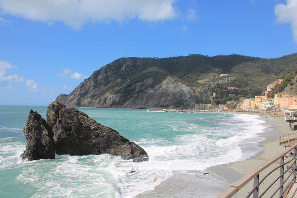 Waves breaking around a dark sea rock beside a curving beach and cliffside houses