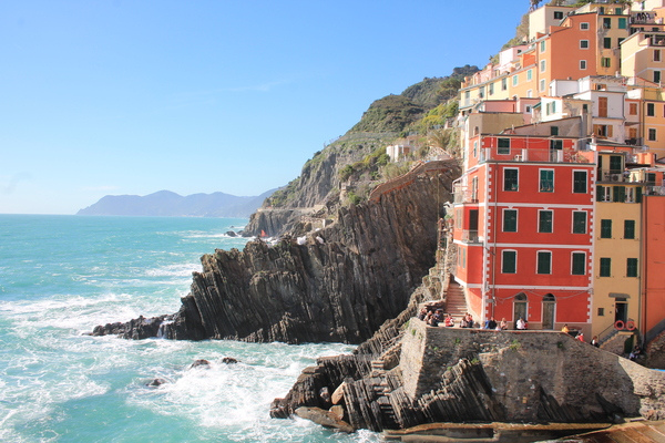 Red and yellow houses perched on layered rocks above a rough turquoise sea in a Cinque Terre village