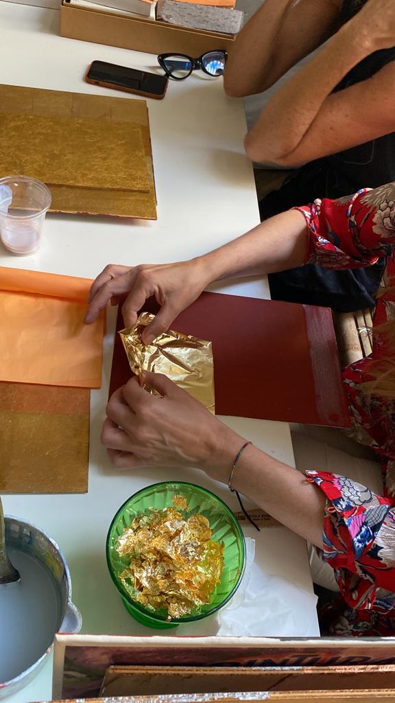 Hands handling gold leaf sheets and flakes at a table during a gilding workshop