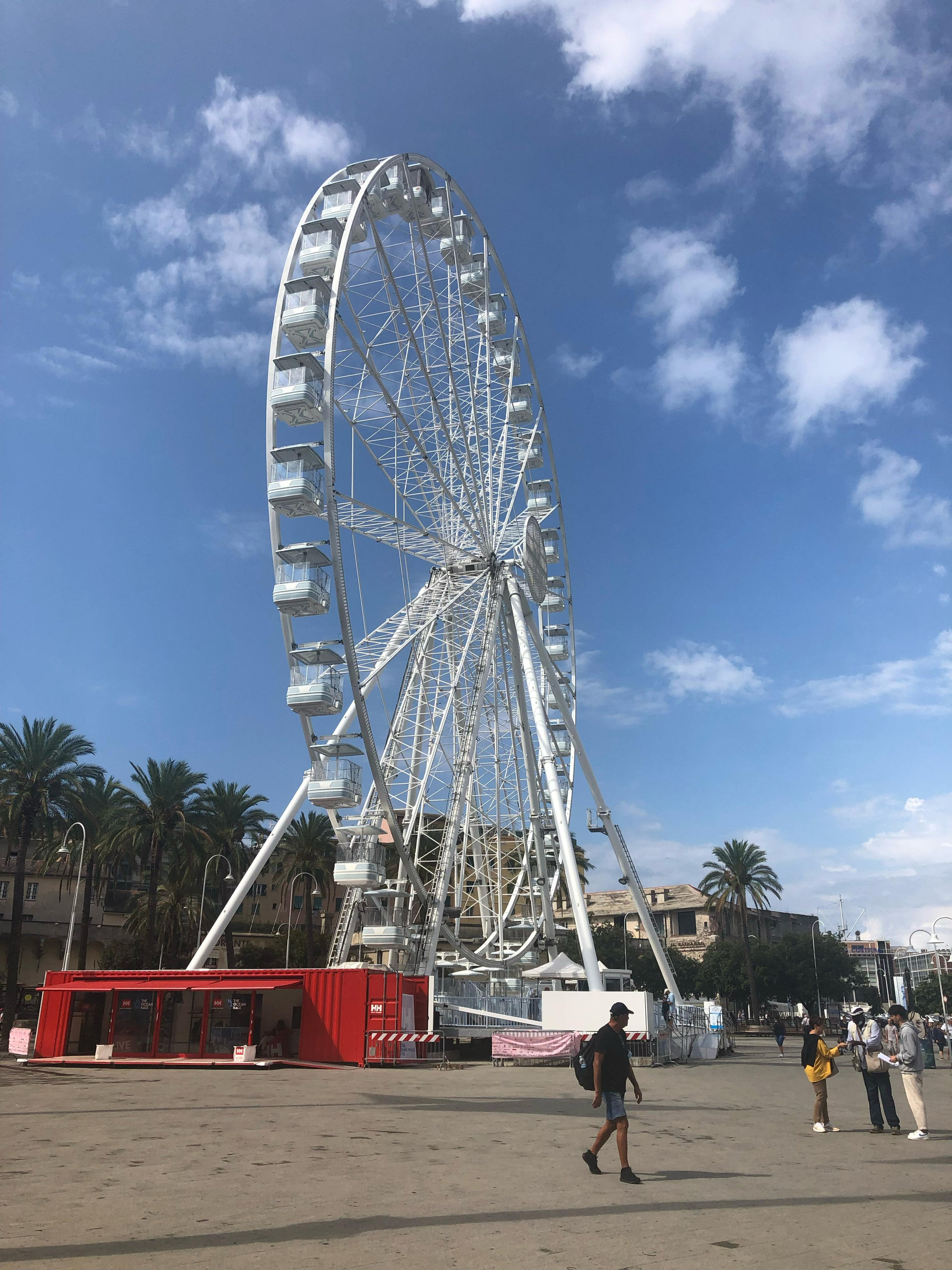 Large white Ferris wheel on a waterfront square with palm trees and passers-by