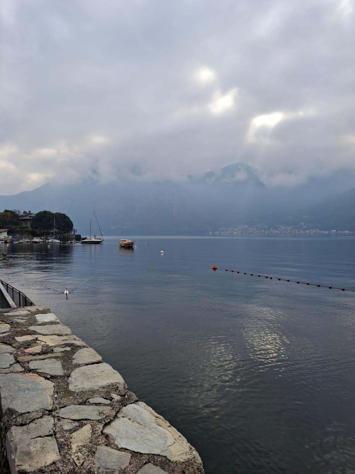 Stone lakeside promenade curving beside still water, with moored boats and misty mountains