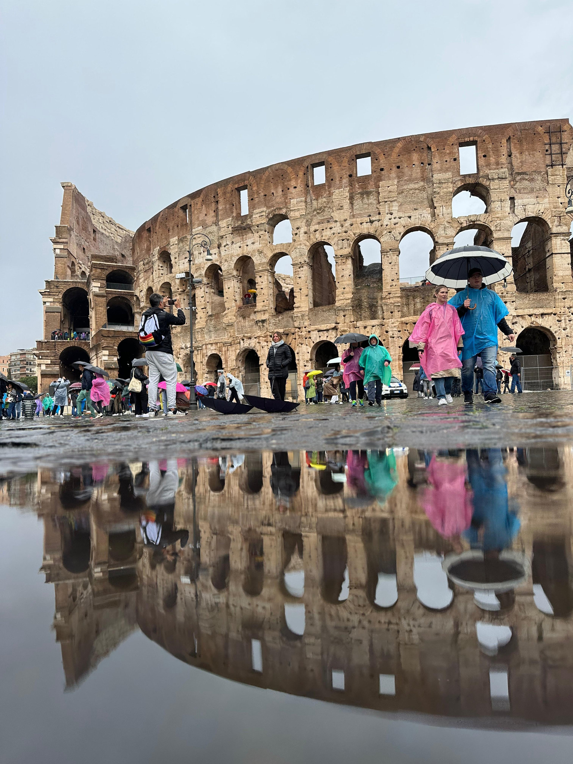 Colosseum reflected in a rain puddle, with visitors in colourful ponchos and umbrellas