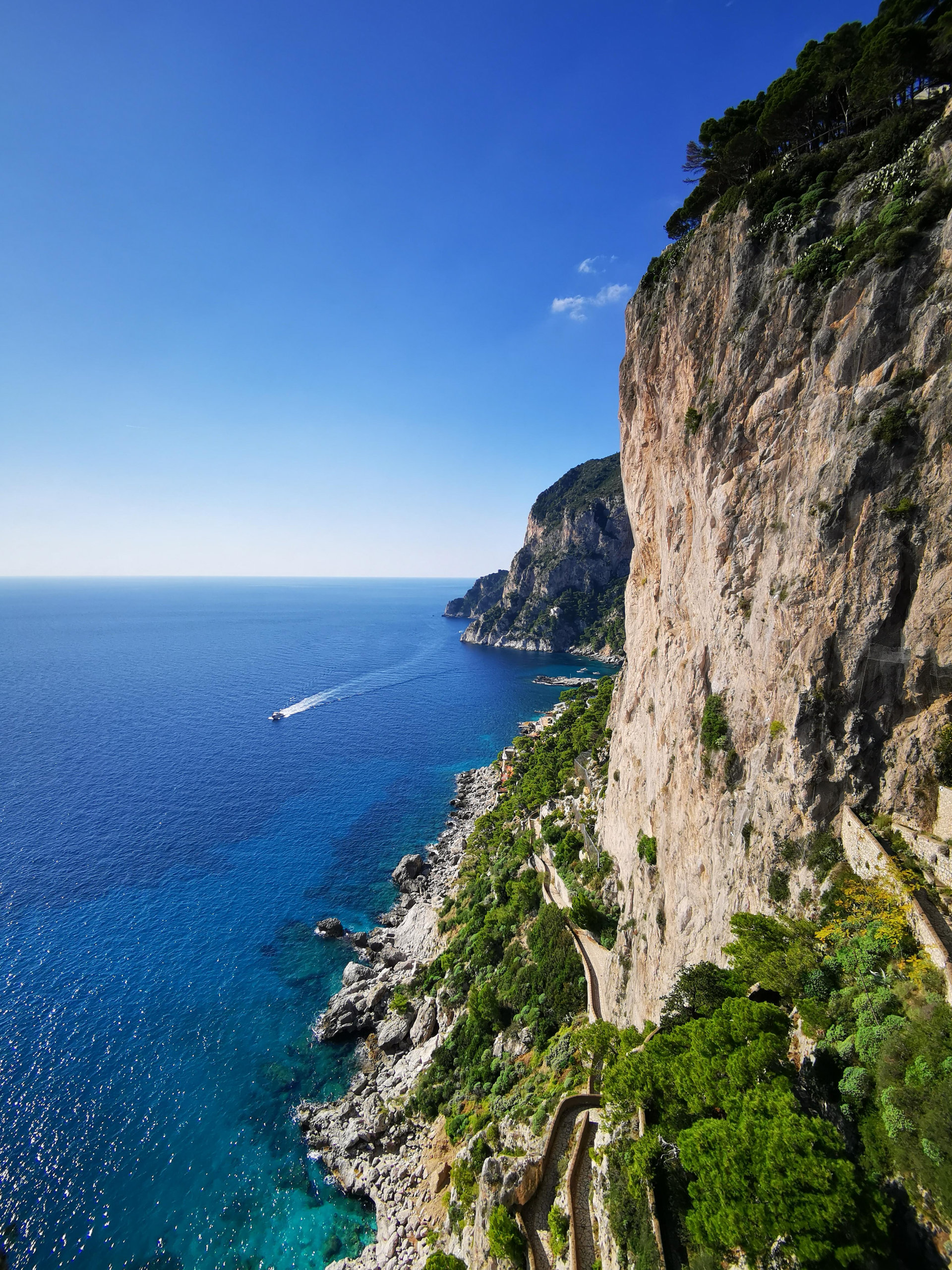 Steep limestone cliff dropping to a turquoise sea, with a path winding down and a boat wake offshore